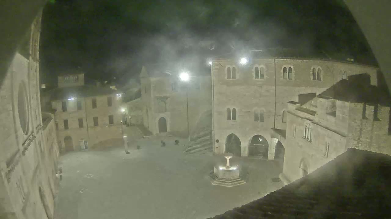 A hazy, nighttime view of a town square with several stone buildings surrounding a central fountain.