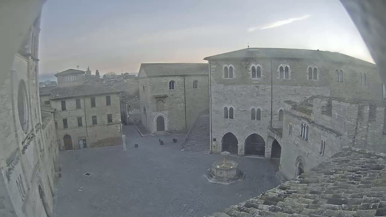 A high-angle, long shot of a town square with several stone buildings surrounding it, a fountain in the center, and a mostly clear, hazy sky.