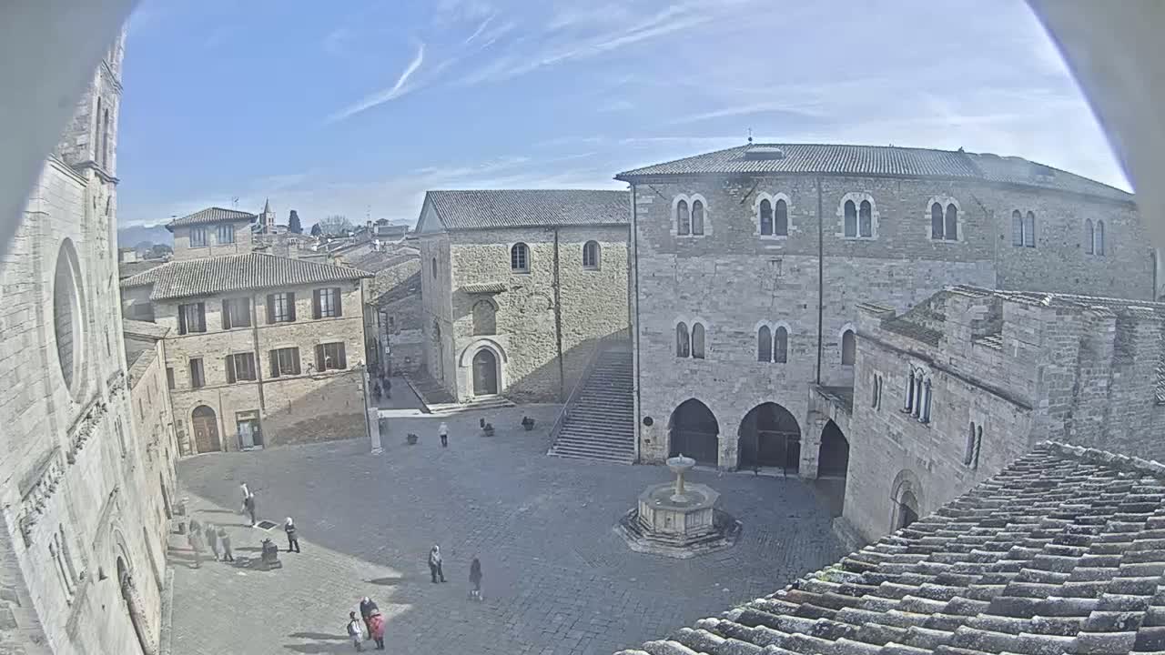 A high-angle view shows a town square in sunny weather, surrounded by ancient stone buildings, one of which features a prominent staircase and fountain.