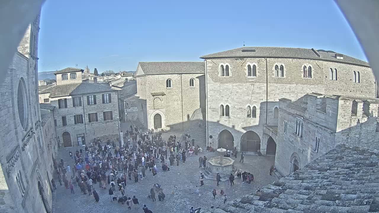 A crowd of people gathers in a stone plaza surrounded by medieval-style buildings on a sunny day.