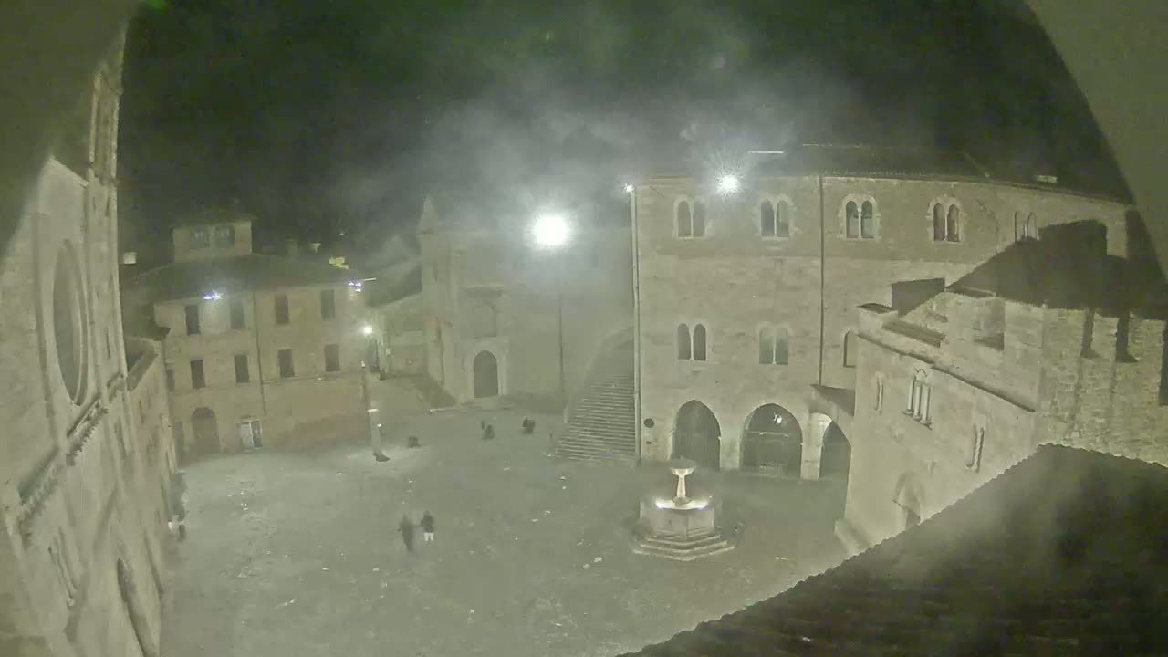 A nighttime view of a town square with stone buildings and a fountain, under hazy or foggy conditions.