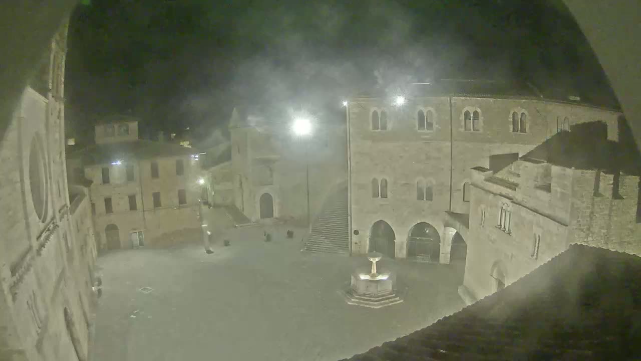 A nighttime view of a town square, partially obscured by fog or mist, shows several stone buildings surrounding a central fountain.