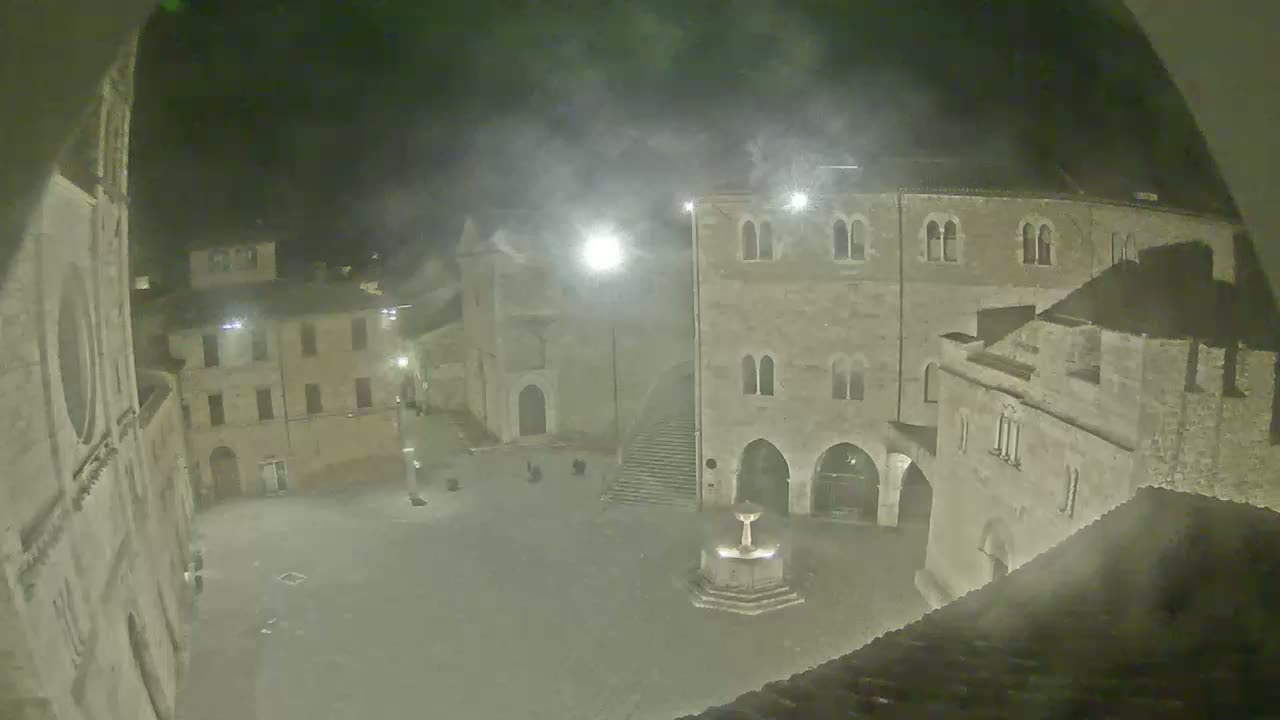 A hazy night scene shows a town square with several stone buildings, a fountain, and a few people in the distance.