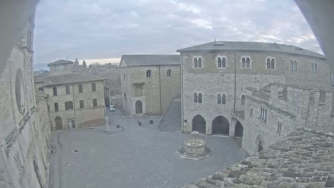 A stone plaza with a fountain is surrounded by medieval-style buildings under an overcast sky.