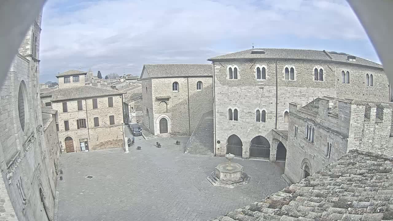 A mostly empty town square, under a partly cloudy sky, is surrounded by aged stone buildings, one featuring a central fountain.