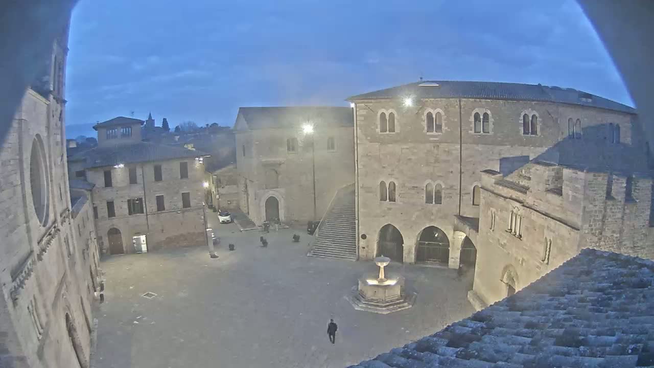 A stone town square at twilight, featuring a fountain, several buildings, and a single pedestrian.
