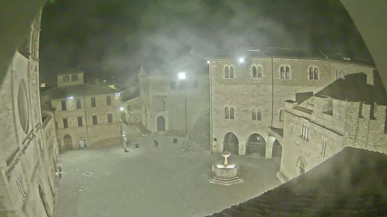 A nighttime view of a town square with stone buildings, a fountain, and a hazy atmosphere.