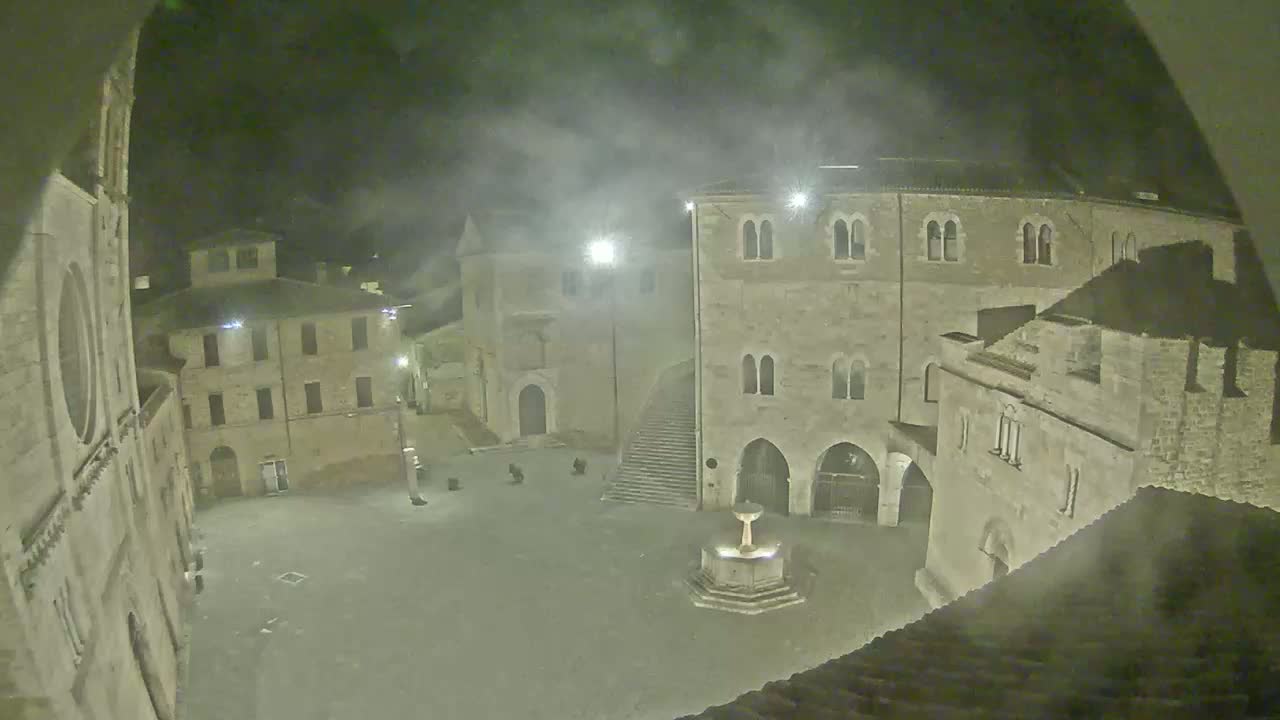 A hazy night scene shows an empty town square with ancient stone buildings surrounding it, one featuring a fountain in the center.