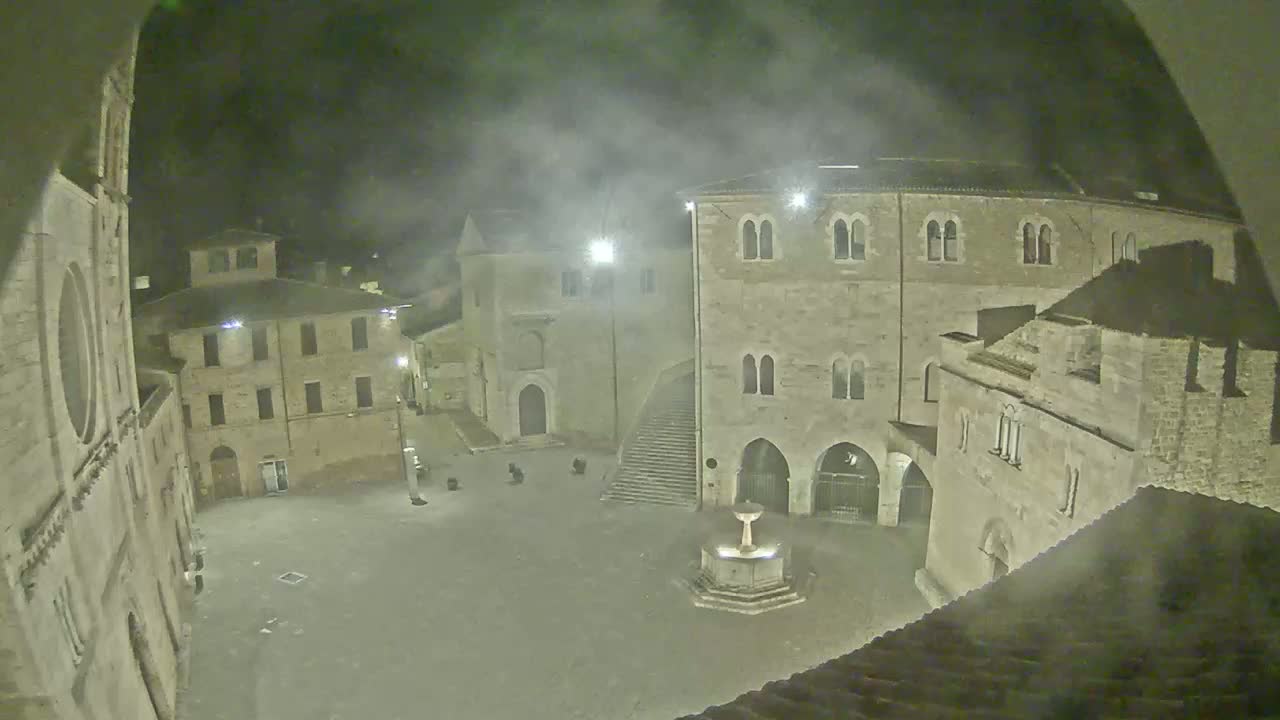 A nighttime view of a town square, partially obscured by fog, shows several stone buildings surrounding a central fountain.