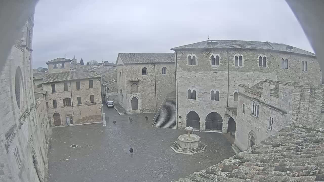 A stone plaza with a fountain is surrounded by medieval-style buildings under an overcast sky.