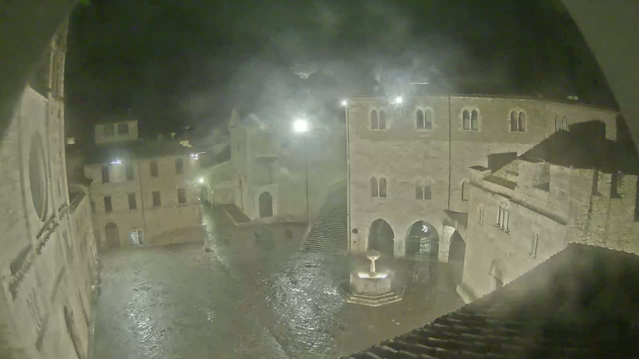 A rain-soaked town square at night, surrounded by old stone buildings, with a fountain at its center.