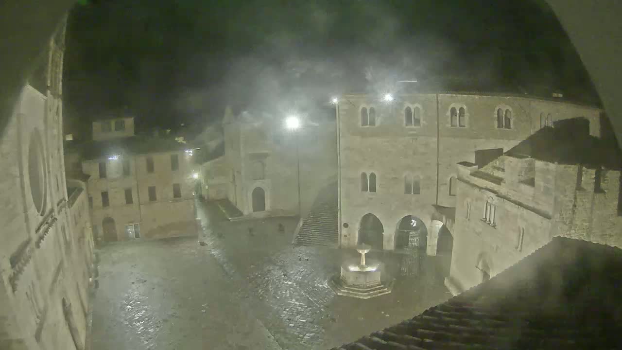 A nighttime view of a town square, showing several stone buildings surrounding a fountain under misty or foggy conditions.