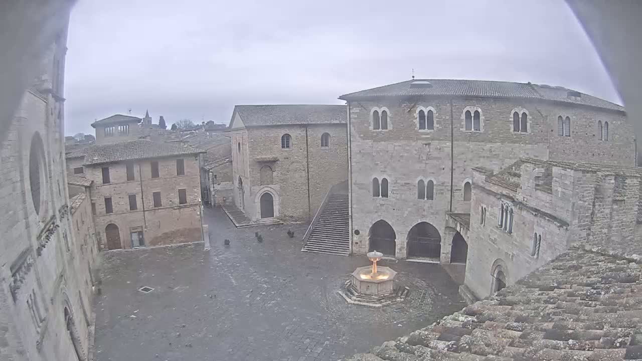 A stone plaza with a fountain is surrounded by medieval buildings under an overcast sky.
