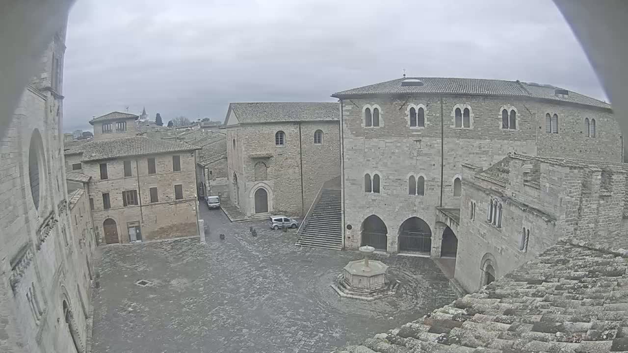 A stone plaza with a fountain is surrounded by several aged stone buildings under an overcast sky.
