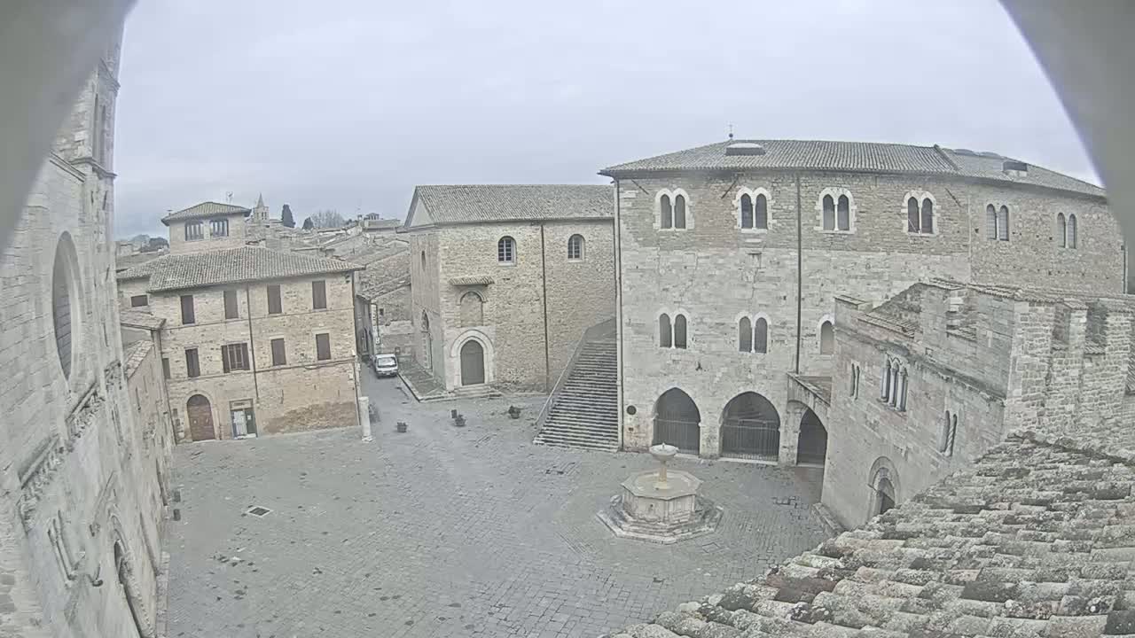 A stone plaza with a fountain is surrounded by medieval-style buildings under an overcast sky.