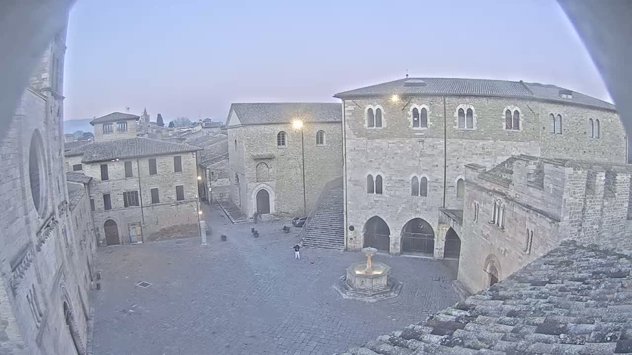 A mostly empty, stone plaza with a fountain is surrounded by ancient-looking buildings under a clear, pale sky.