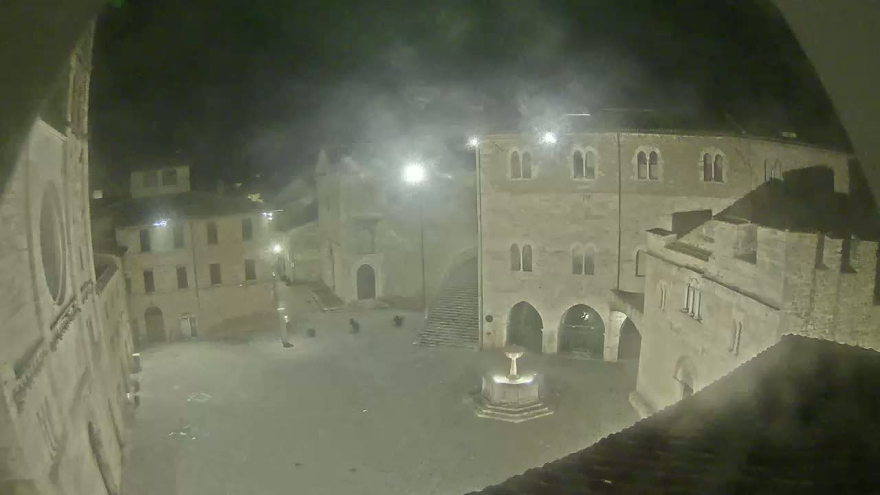 A nighttime, hazy view of a town square with old stone buildings surrounding a fountain.