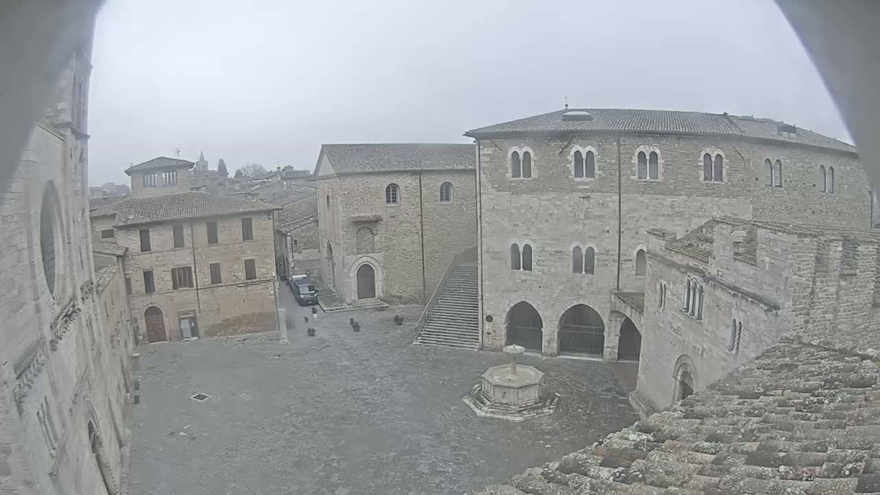 A stone town square with several medieval-style buildings, a fountain, and a few people, viewed on an overcast day.