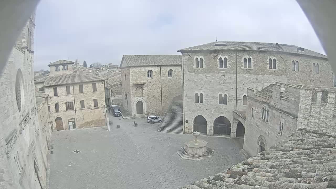 A mostly empty cobblestone square in a historic town is surrounded by aged stone buildings under an overcast sky.