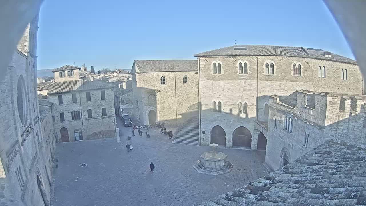 A high-angle view of a town square on a sunny day, featuring several stone buildings surrounding a central fountain and a few people walking around.