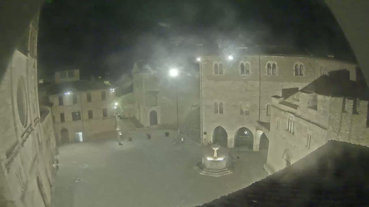 A nighttime, hazy view of a town square with stone buildings surrounding a central fountain.