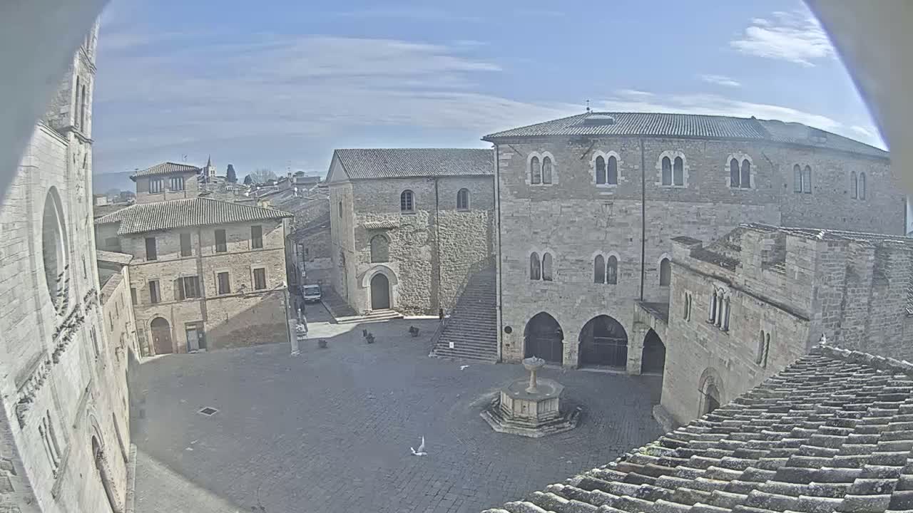 A cobblestone town square, under a mostly sunny sky, is surrounded by historic stone buildings, one featuring a fountain at its center.