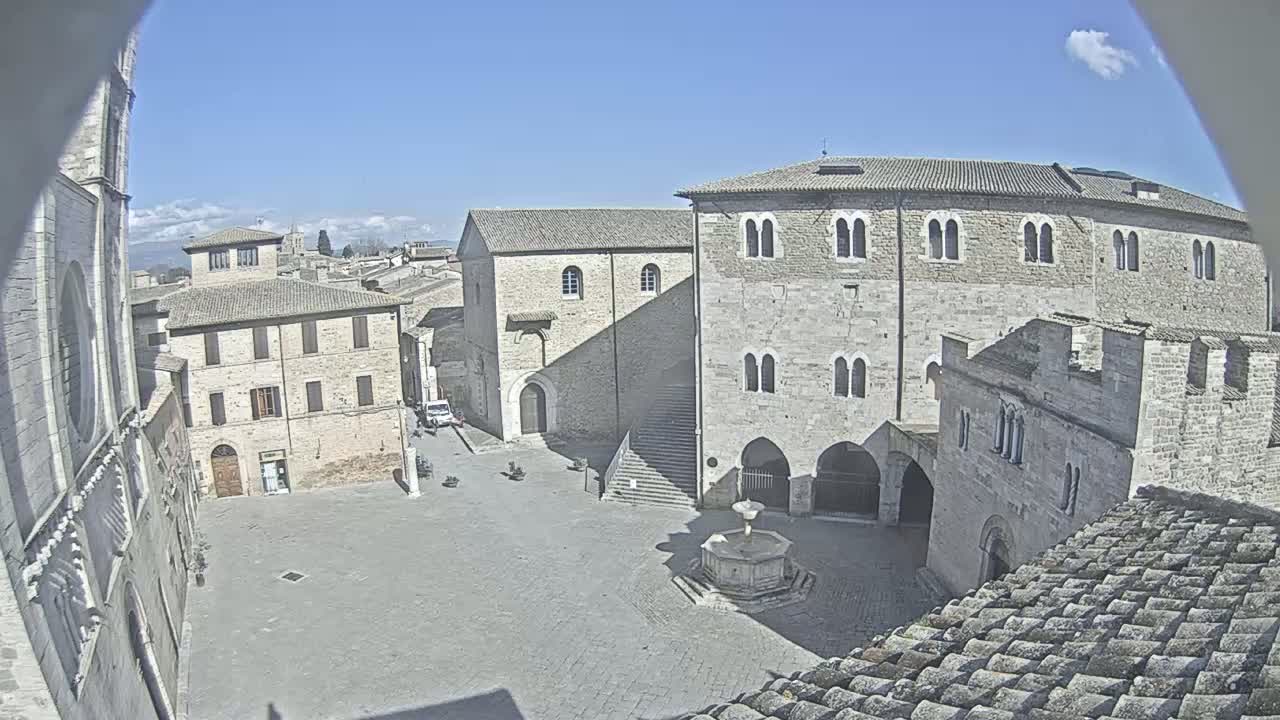 A paved town square with a fountain, surrounded by ancient stone buildings, under a clear blue sky.