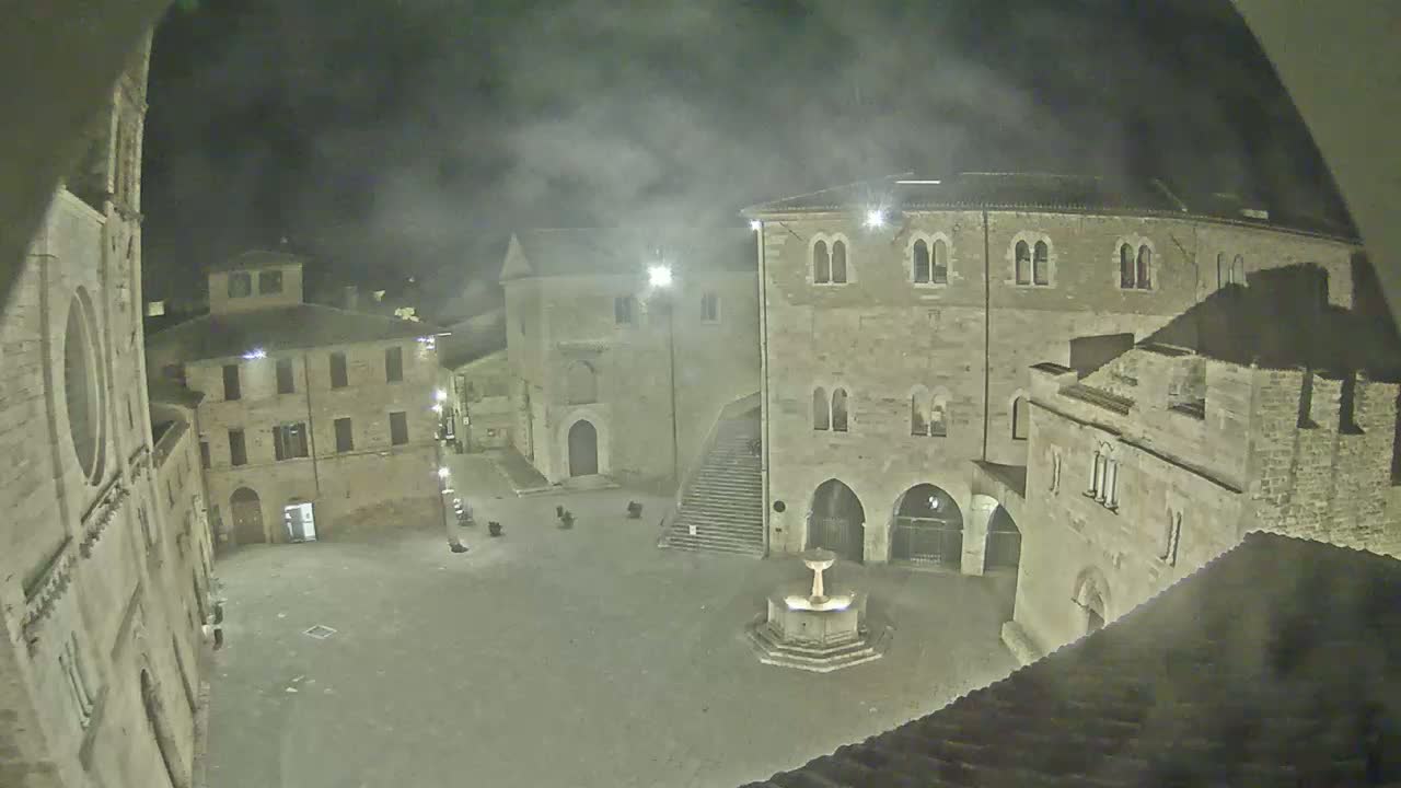 A nighttime view of a stone plaza with a fountain, surrounded by old buildings under hazy or smoky conditions.