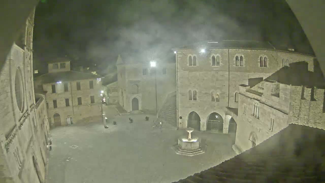 A nighttime view of a stone plaza with a fountain, surrounded by ancient buildings under hazy or foggy conditions.
