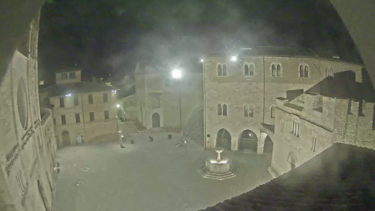 A nighttime view of a stone plaza with a fountain, surrounded by old buildings under hazy or foggy conditions.