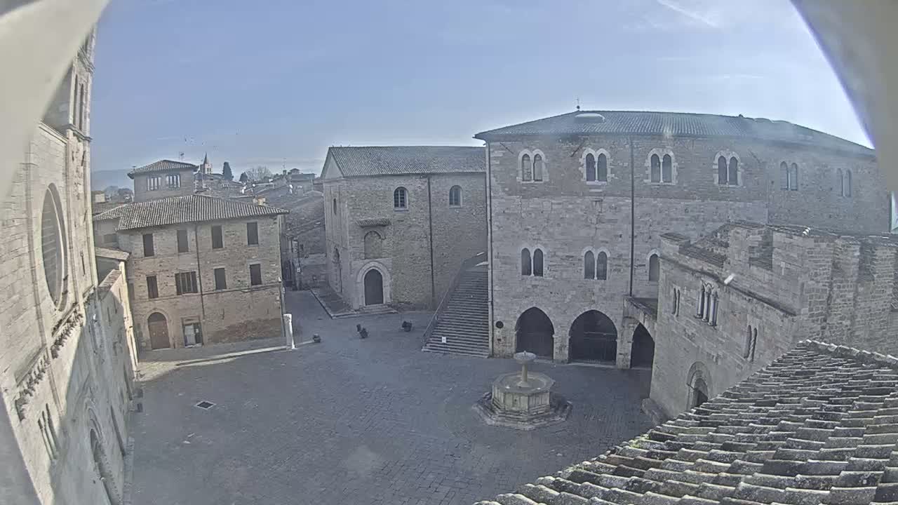 A stone plaza with a fountain, surrounded by medieval-style buildings, on a clear sunny day.