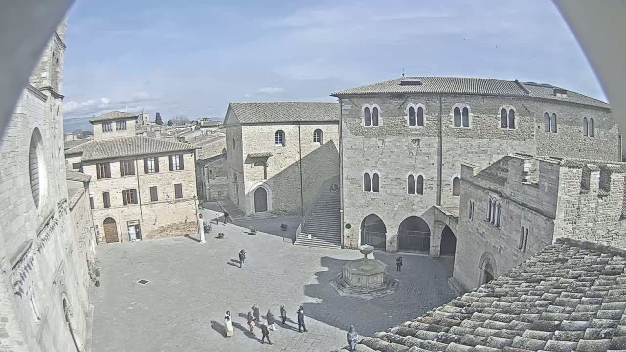 A mostly empty town square, under a clear sunny sky, is surrounded by ancient stone buildings, including a large, imposing structure with arched entryways and a smaller building with a visible staircase.