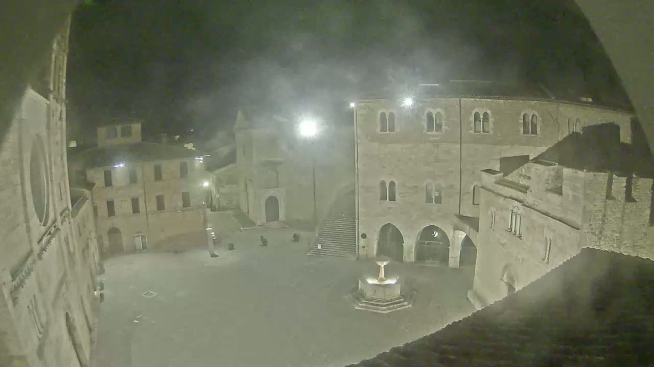 A nighttime view of a town square with stone buildings, a fountain, and hazy weather conditions.