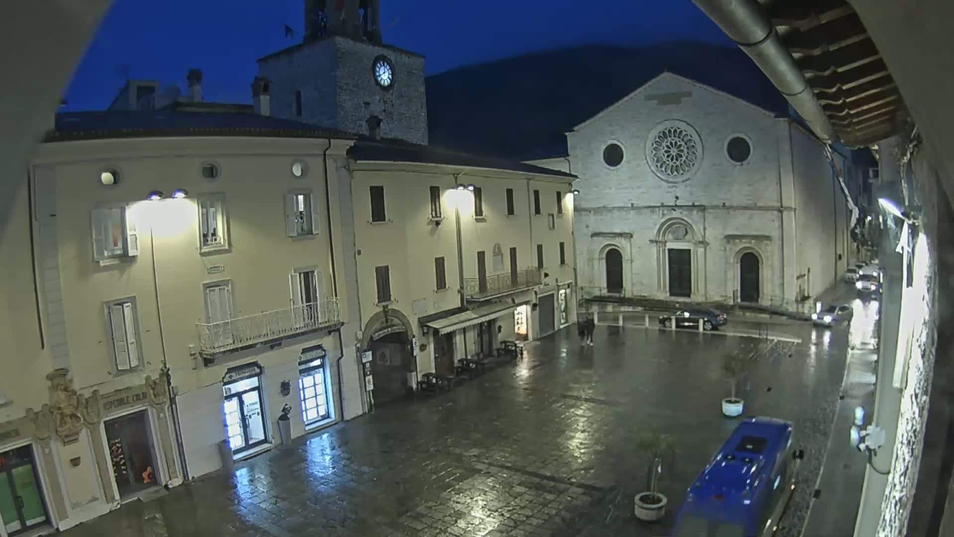 A rain-slicked town square at night, illuminated by streetlights, features several buildings including a clock tower and a church with a rose window.