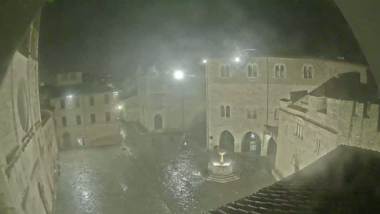 A town square is flooded at night under heavy rain, with old stone buildings surrounding the waterlogged area.