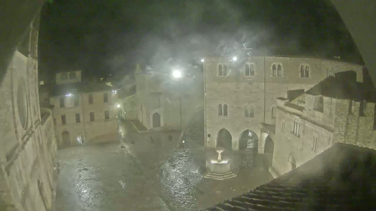 A rain-swept town square at night, surrounded by old stone buildings, features a fountain in the center.