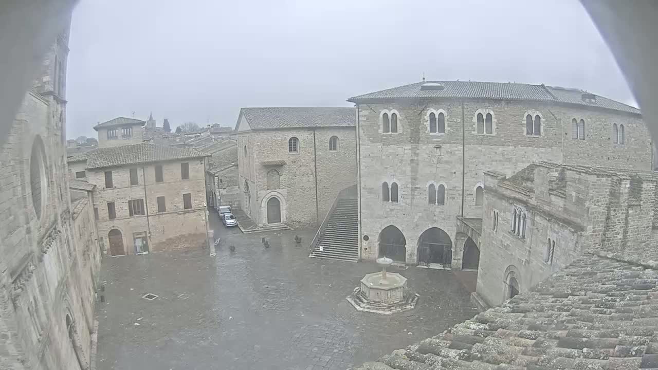 A stone plaza with a fountain is surrounded by medieval-looking buildings under an overcast sky.
