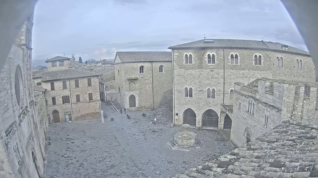 A stone plaza, under an overcast sky, is surrounded by medieval-style buildings, one of which has a prominent staircase.