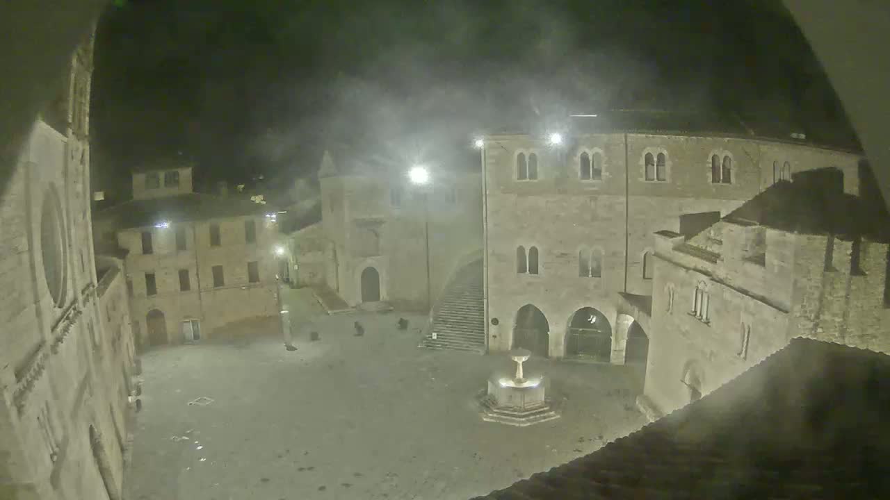 A nighttime view of a town square, partially obscured by mist or fog, shows several stone buildings surrounding a fountain.