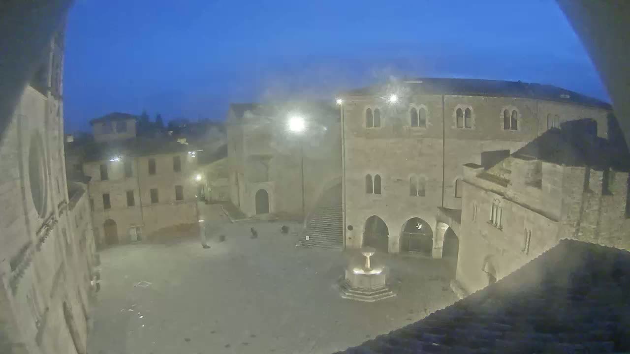 An empty town square at night, under a dark blue, possibly hazy sky, features a fountain and several stone buildings surrounding it.