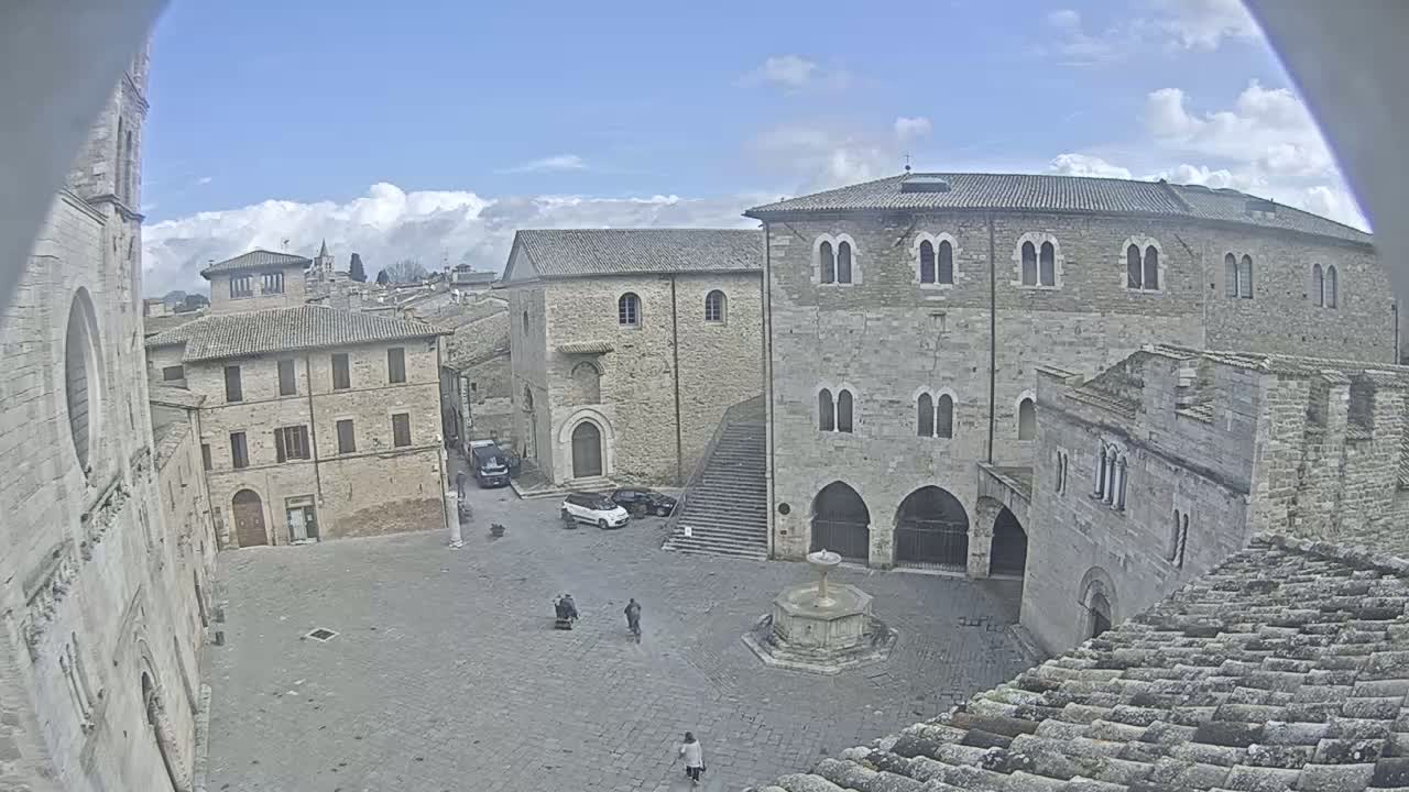 A stone town square with a fountain, surrounded by old buildings, under a partly cloudy sky.