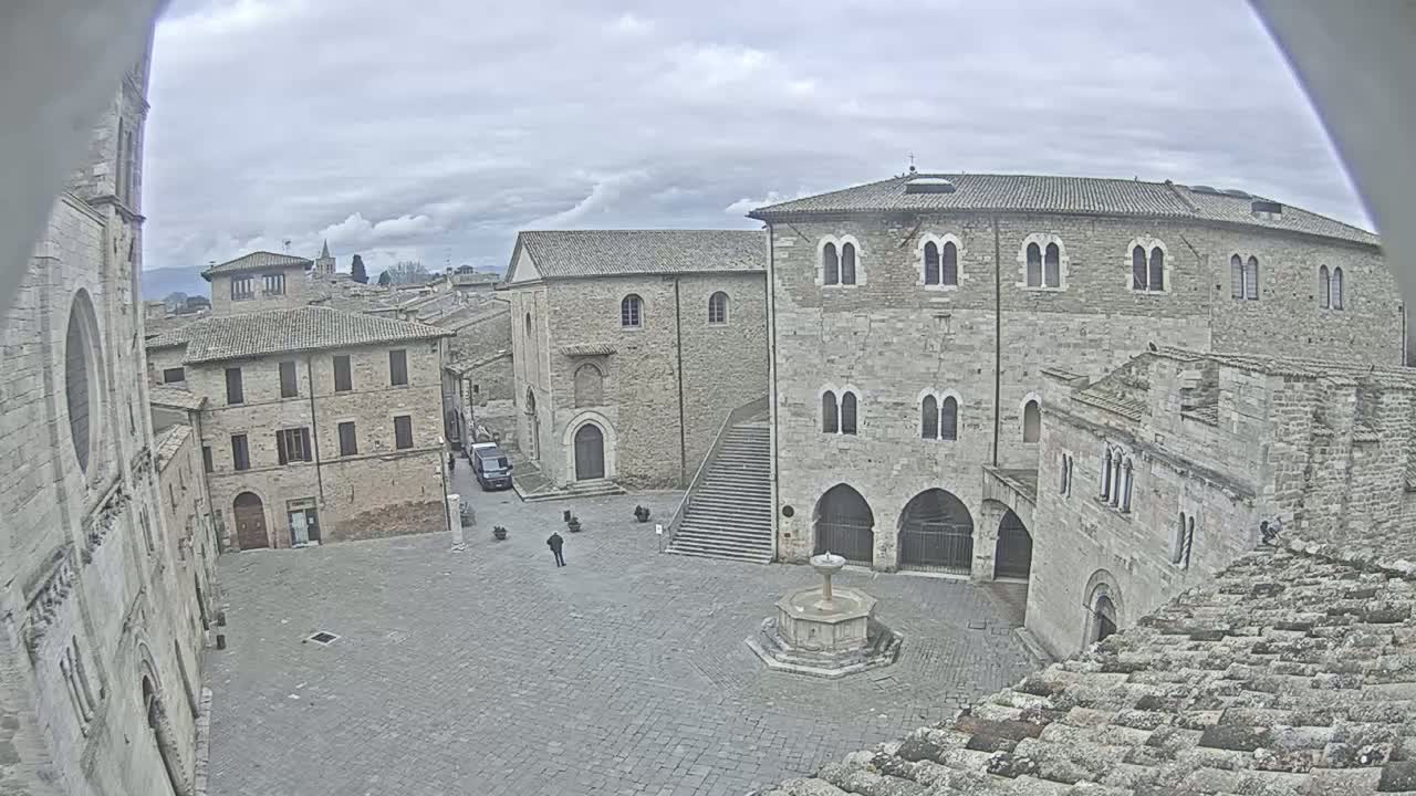 A stone plaza with a fountain is surrounded by medieval-style buildings under a cloudy sky.