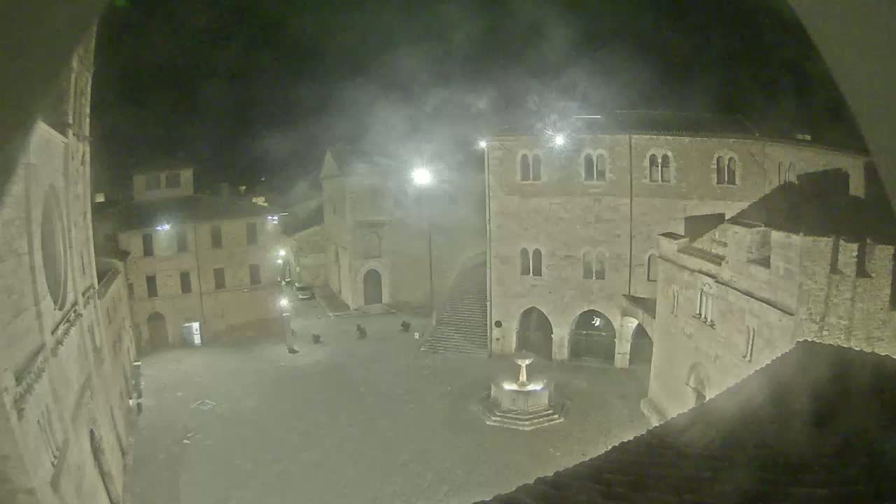 A nighttime view of a town square with a fountain, surrounded by stone buildings under hazy conditions.