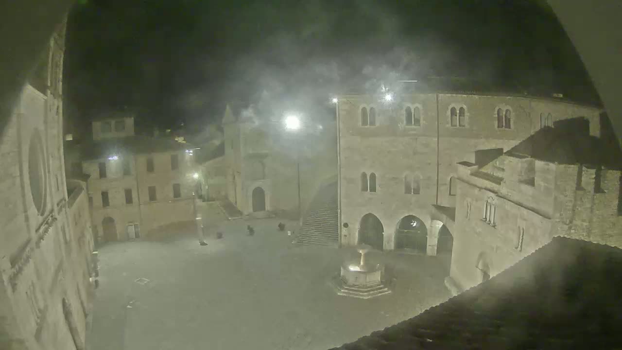 A nighttime view of a town square, shrouded in fog or mist, features several historic stone buildings surrounding a central fountain.