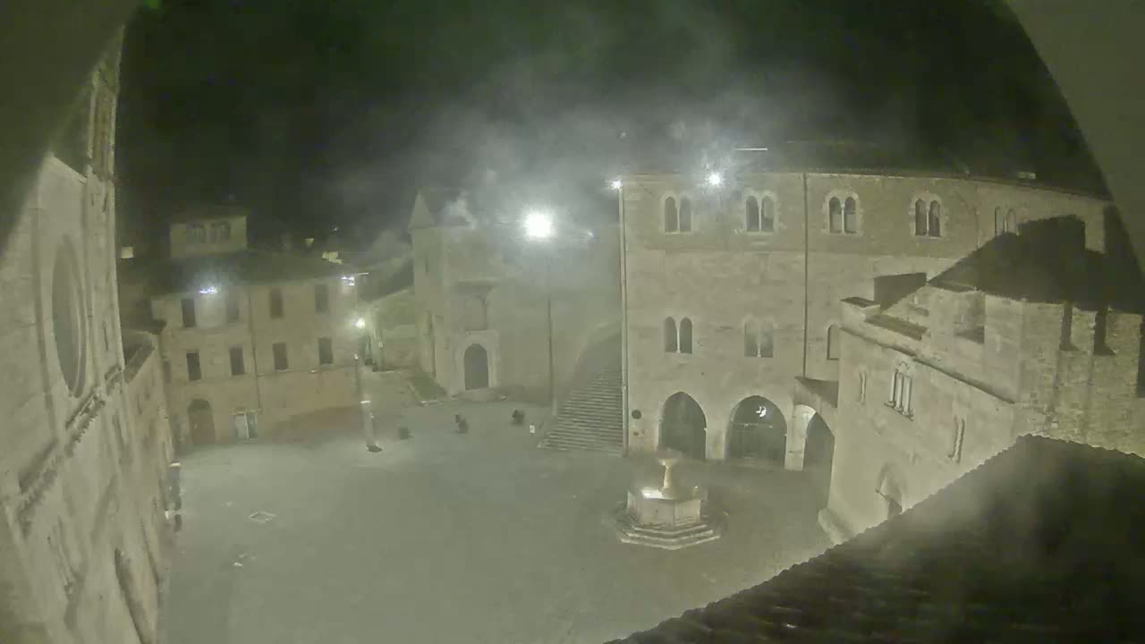 A nighttime, hazy view of a town square with stone buildings surrounding a central fountain.