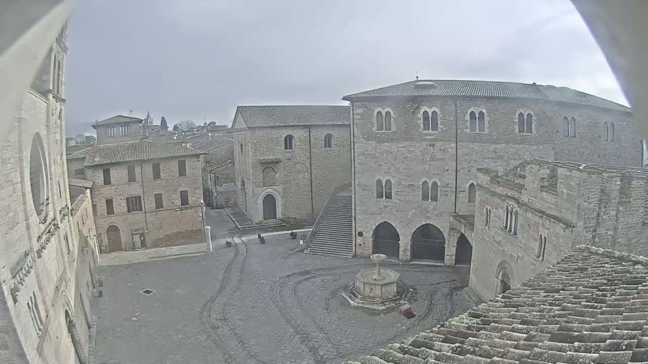A stone plaza with several buildings, a fountain, and a staircase under an overcast sky.