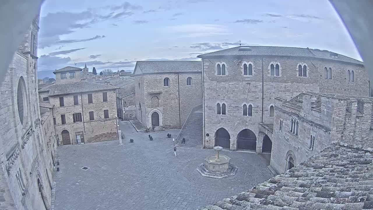 A stone plaza with a fountain is surrounded by medieval-style buildings under a mostly cloudy sky.