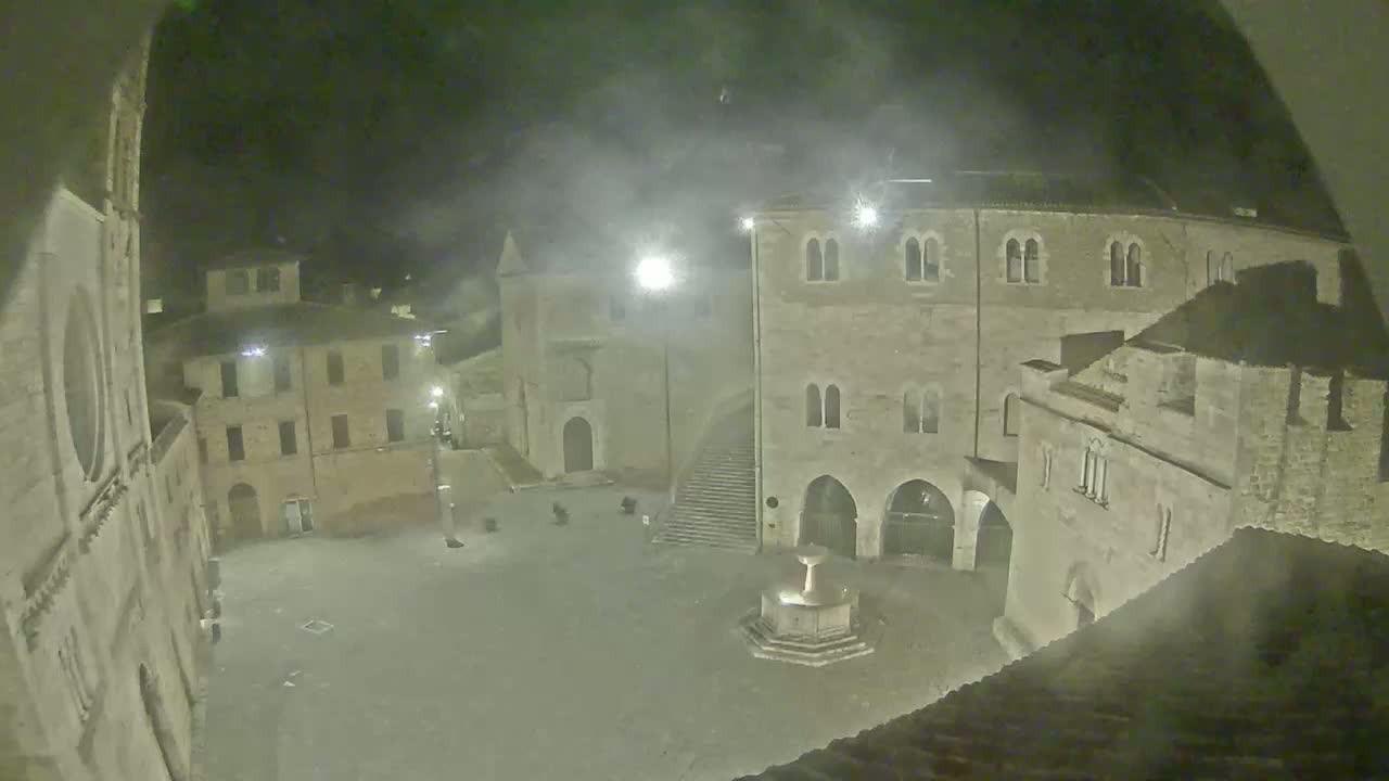 A nighttime, hazy view overlooking a town square with aged stone buildings surrounding a central fountain.