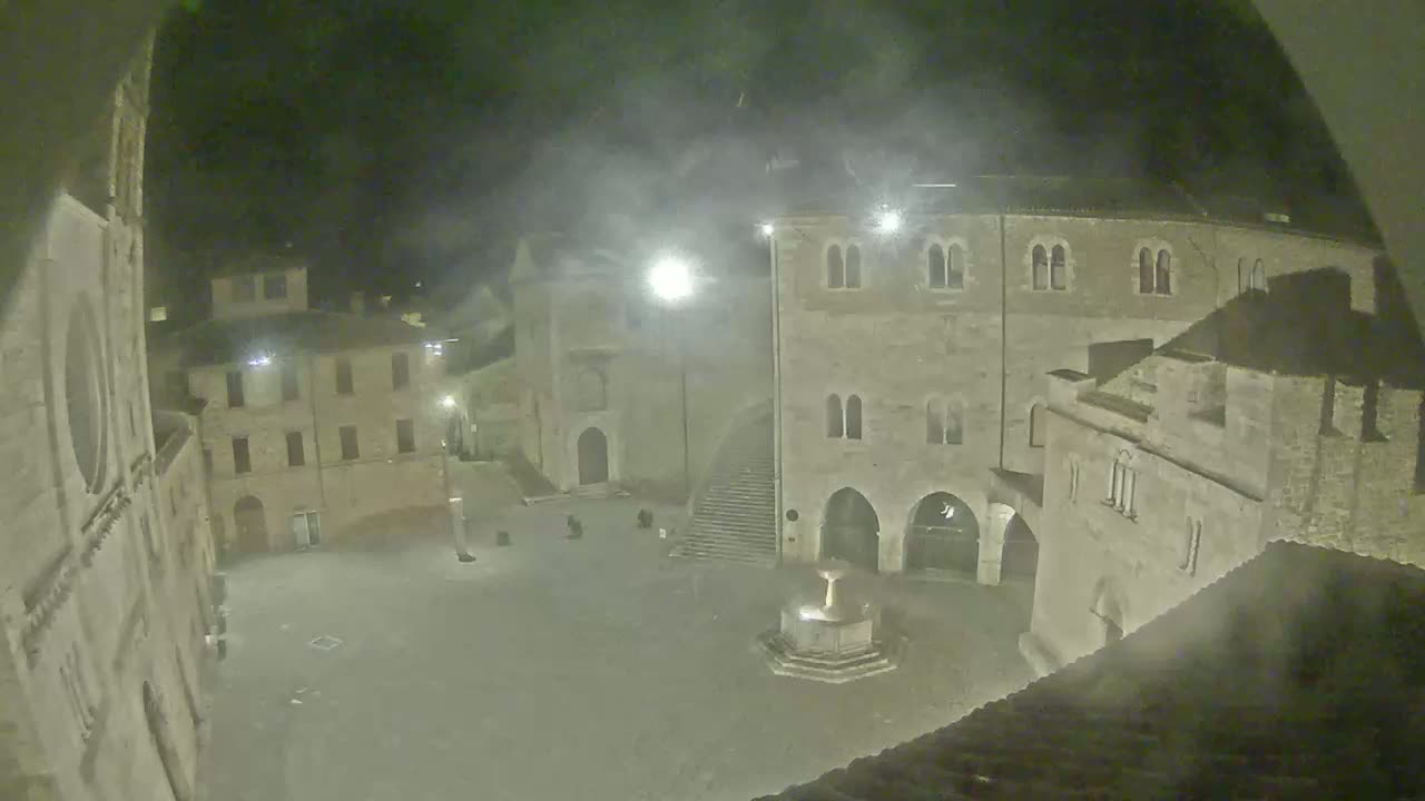 A nighttime view of a town square, shrouded in mist or fog, features ancient stone buildings surrounding a central fountain.