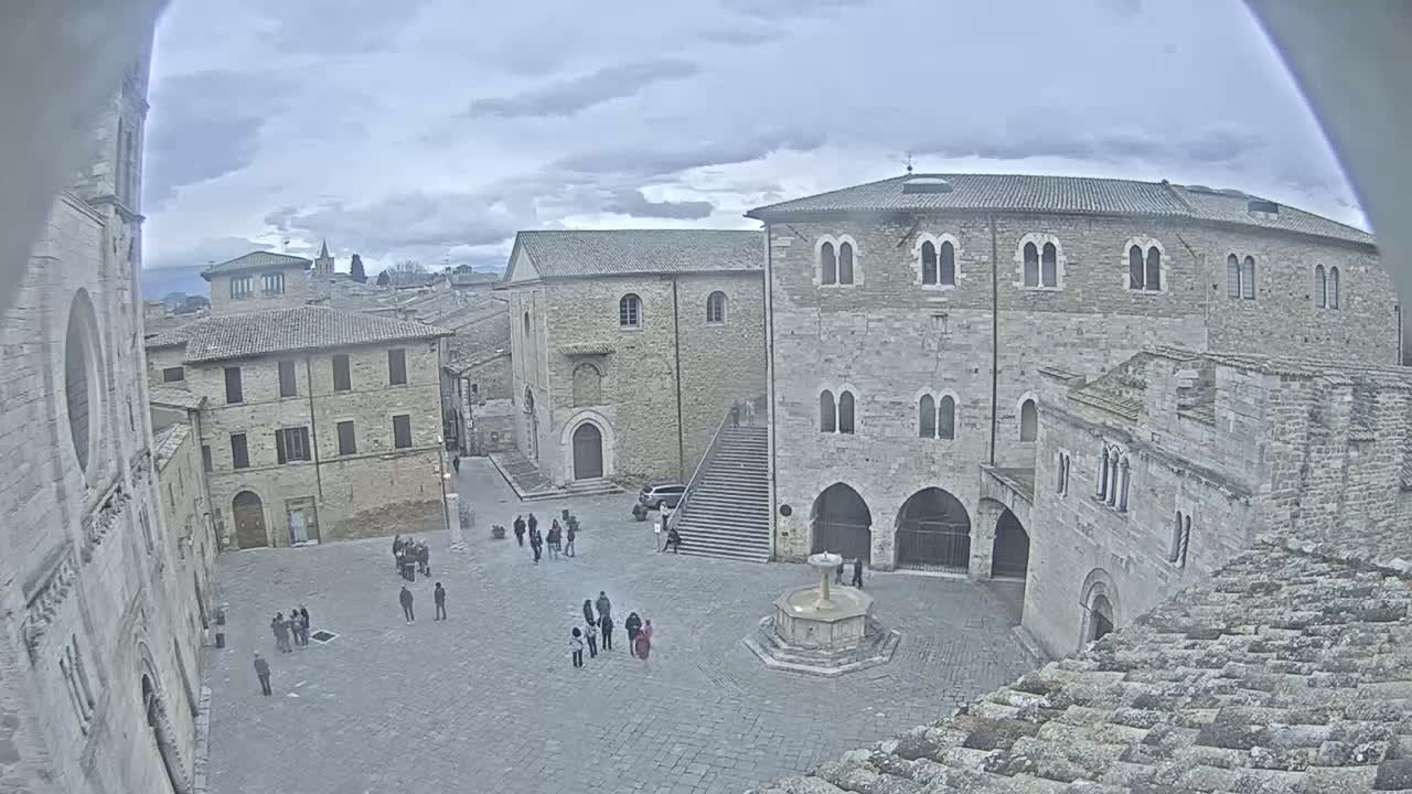 An overcast day reveals a high-angle view of a town square, centered around a fountain, with several stone buildings and a scattering of people.